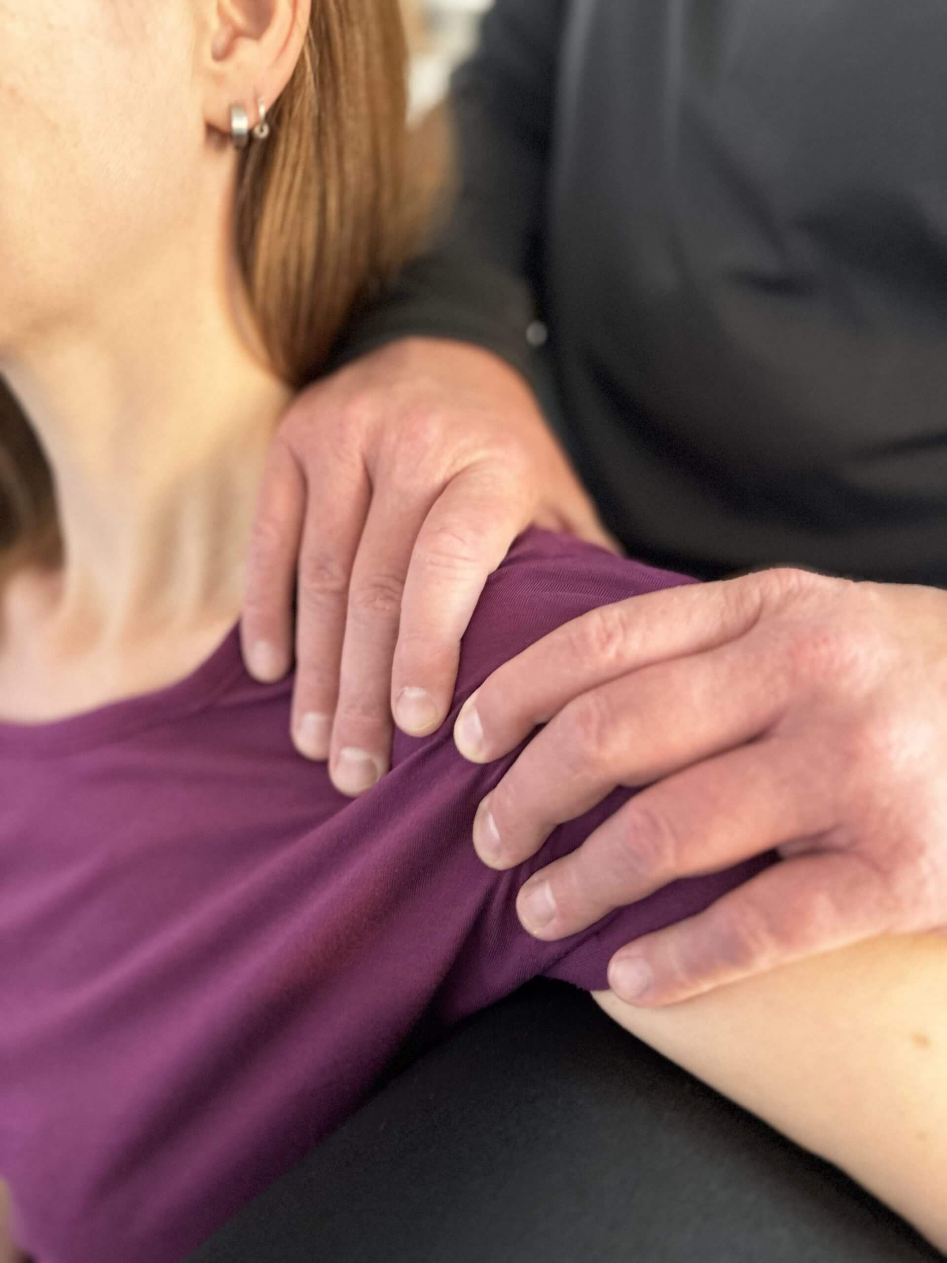 Physiotherapeut behandelt mit beiden Händen die Schulter einer sitzenden Patientin