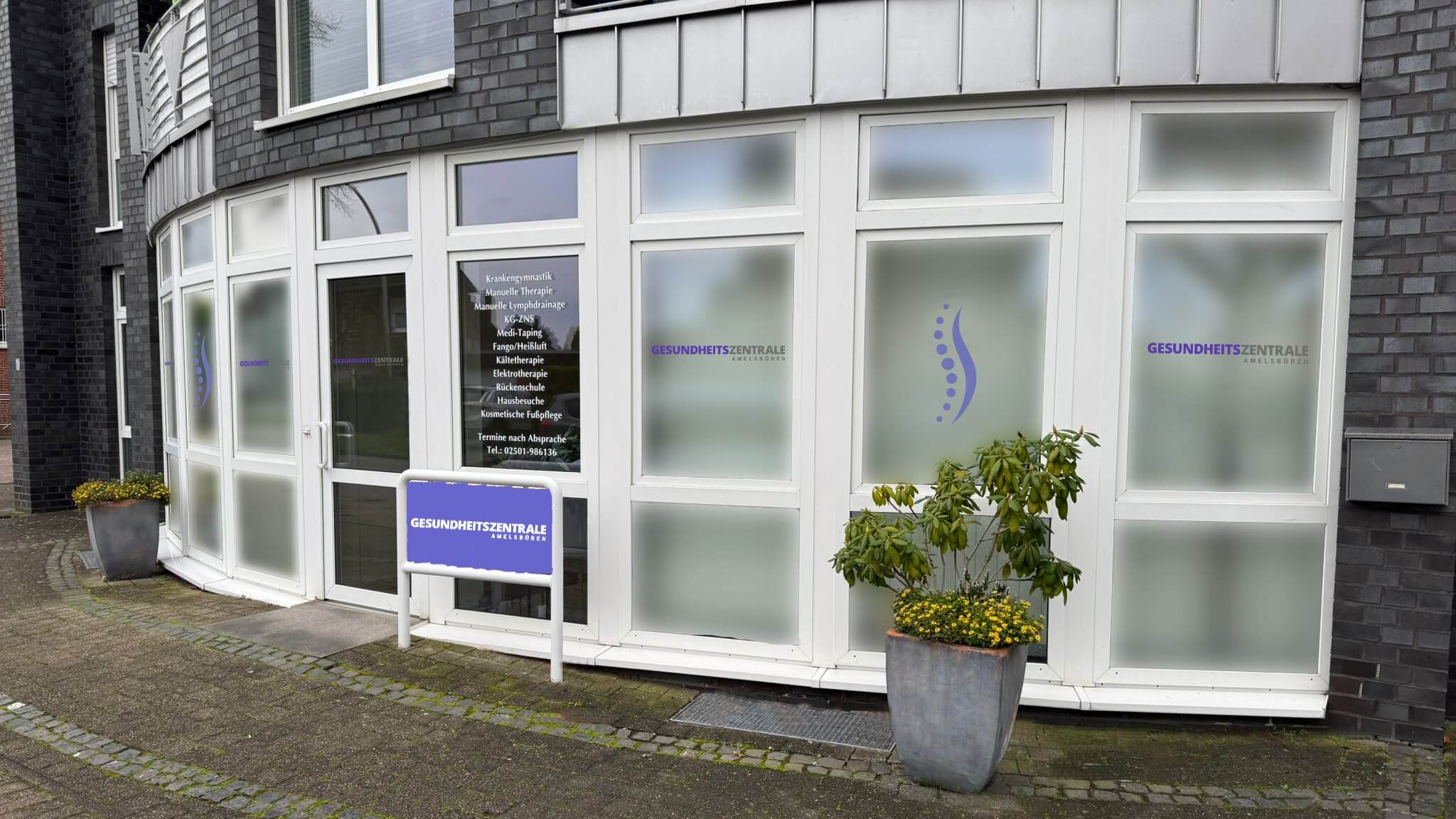 Eingang der Gesundheitszentrale Amelsbüren mit Praxisschild, Logo auf den Fenstern und Leistungsübersicht an der Tür